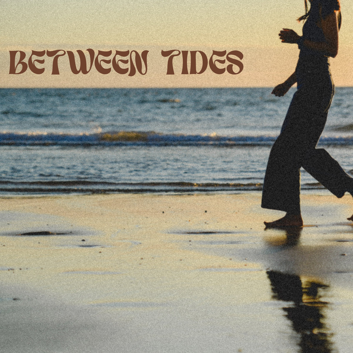 A woman walks barefoot along a wet sandy beach at sunset, with the ocean and sky behind her. The words 'BETWEEN TIDES' appear across the top in a stylized brown font.